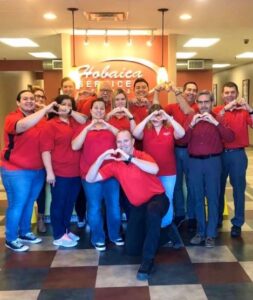 A group of people in red shirts stand indoors forming heart shapes with their hands, smiling at the camera. The sign behind them reads "Hobaica Service.
