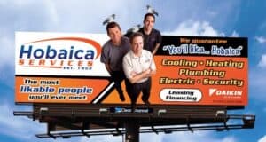 Billboard for Hobaica Services featuring three men smiling, two with pigeons on their heads. Text advertises cooling, heating, plumbing, electric, security services, and leasing/financing options.
