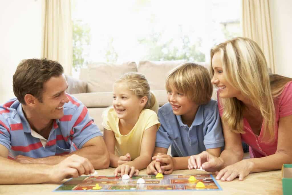 Four people, two adults and two children, are lying on the floor together, smiling and playing a colorful board game in a well-lit living room.