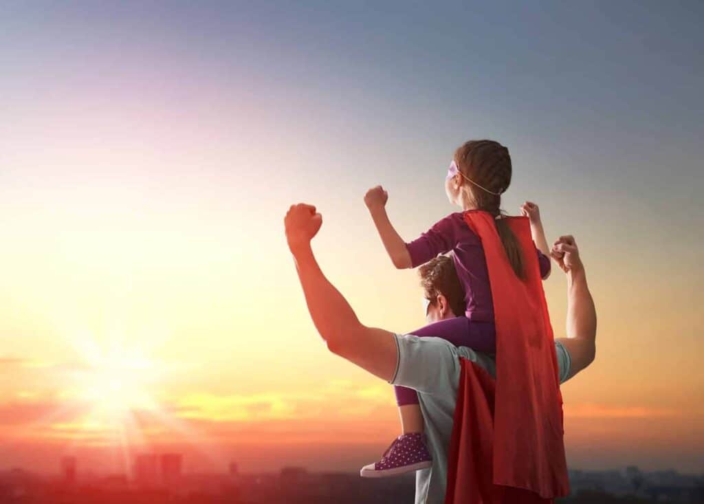 An adult carries a child on their shoulders at sunset. Both wear red capes and raise their fists, looking toward the sky.