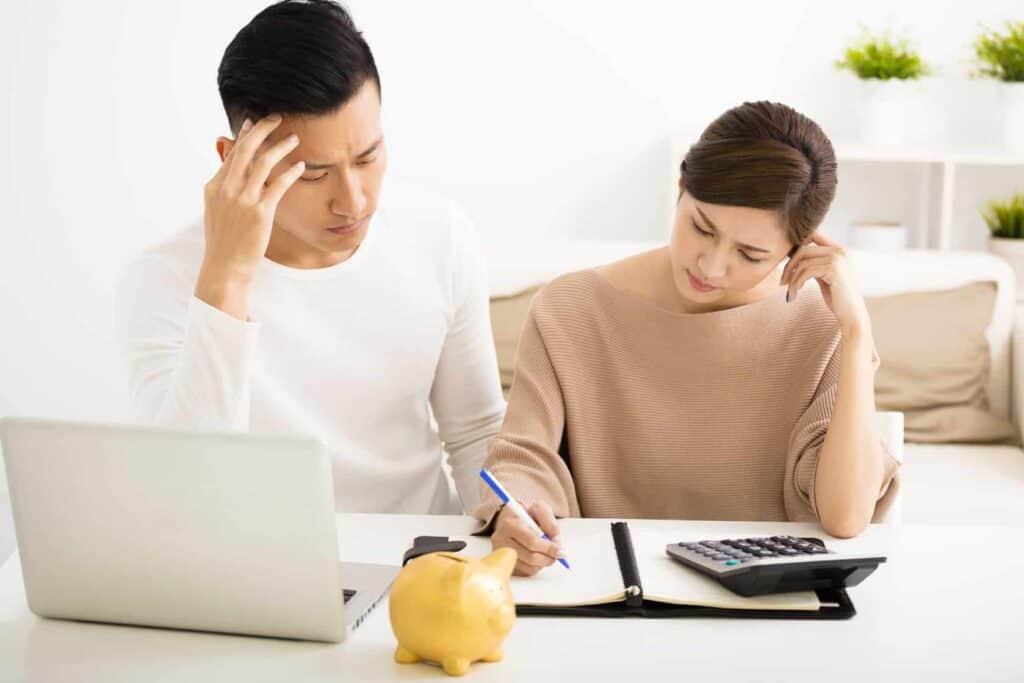 A man and woman sit at a table with a laptop, notebook, calculator, and piggy bank, appearing focused and concerned as they review financial documents.