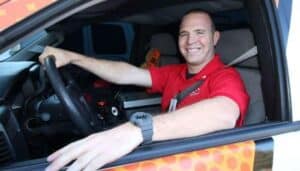 Man in a red shirt and watch sitting in the driver’s seat of a vehicle, smiling at the camera with his left arm resting on the open window.
