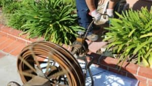A person uses a drain cleaning machine with a spinning cable outdoors near green plants and red brick edging.
