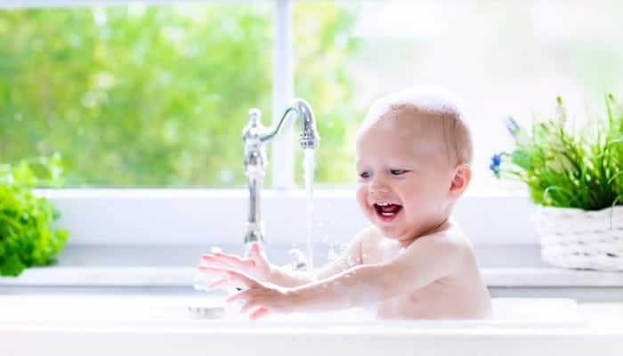 A baby sits in a kitchen sink with running water, smiling and playing with the water. The background shows greenery and a white potted plant near a window.