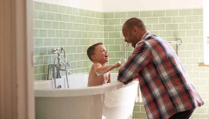 An adult leans over a bathtub talking to a young child who is sitting in the tub and laughing. The bathroom has green tile walls.