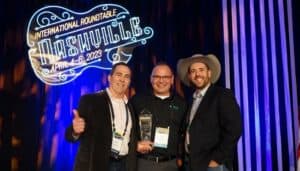 Three men pose smiling with an award in front of a "International Roundtable Nashville April 4-6, 2023" neon sign and striped backdrop.