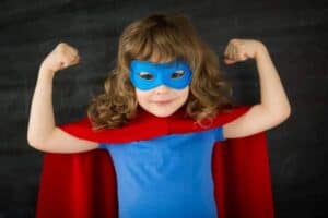 Child in a blue shirt and mask with a red cape stands in front of a chalkboard, smiling and flexing both arms.