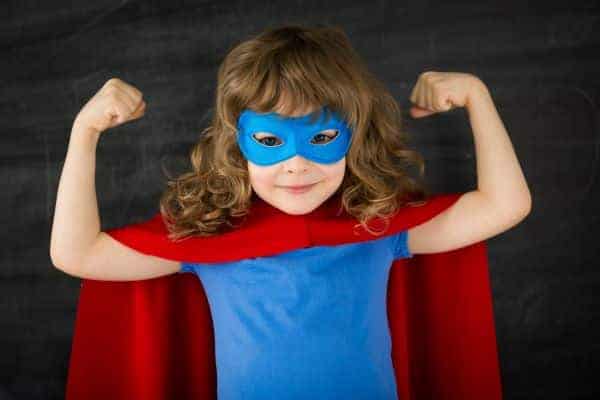 Child in a blue shirt and mask with a red cape stands in front of a chalkboard, smiling and flexing both arms.