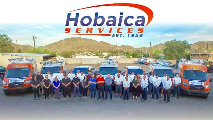 A group of Hobaica Services employees stands in front of company vans, with mountains and buildings in the background and the Hobaica Services logo above.