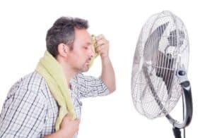 A man with a towel around his neck wipes his forehead while sitting in front of an electric fan.