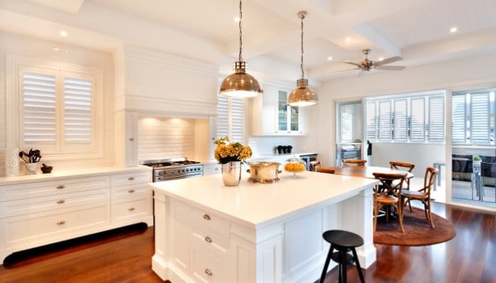 Spacious modern kitchen with white cabinets, central island, pendant lights, stainless steel appliances, wooden dining table, and large windows with shutters.