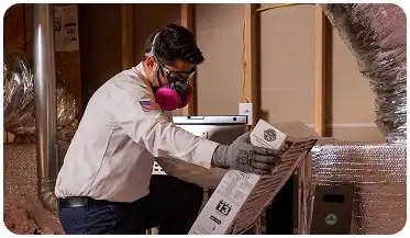 A worker in safety gear installs or inspects an HVAC filter in a utility room with exposed ducts and insulation.