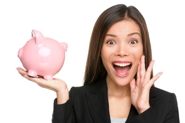 Woman in a black blazer holds a pink piggy bank and looks surprised with her mouth open and hand on her cheek, against a white background.