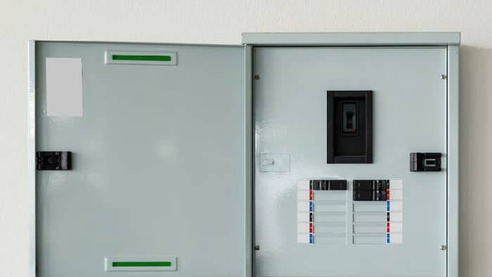 A grey electrical breaker panel with the door open, showing circuit breakers and labels inside, mounted on a white wall.