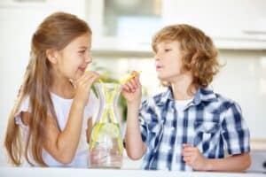 Two children share a glass pitcher of lemon water using yellow straws in a bright kitchen.