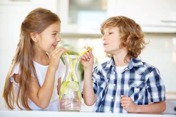 Two children share a glass pitcher of lemon water using yellow straws in a bright kitchen.
