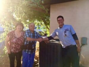 Two people smile while standing beside a service technician who is shaking hands with one of them next to an outdoor HVAC unit.