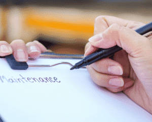 A hand writes "Maintenance" on a paper attached to a clipboard with a black pen.