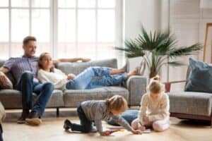 Two children draw on the floor with markers while two adults relax on a gray sofa in a bright living room with large windows and a potted plant.
