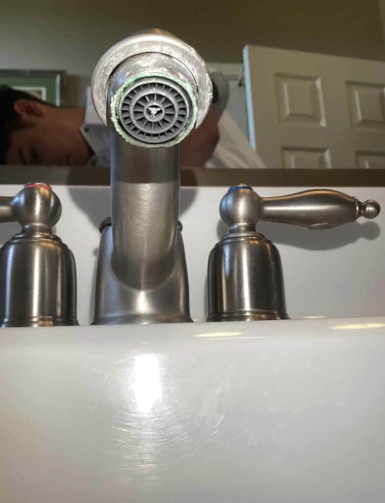 Close-up of a bathroom sink faucet with a person reflected in the mirror behind it. The faucet has mineral buildup around the spout.