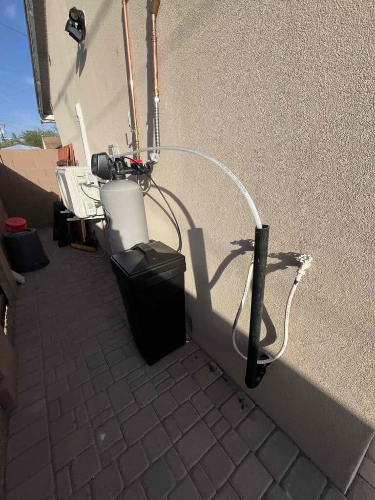 Outdoor water softener system with black brine tank, connected pipes, and cables installed against a stucco wall on a paved walkway in sunlight.