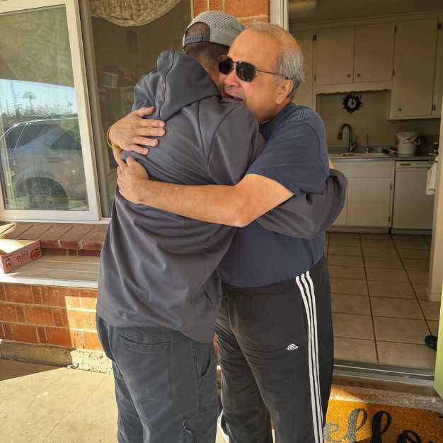 Two men embrace in a hug outside a house, standing near an open door. The kitchen is visible in the background.