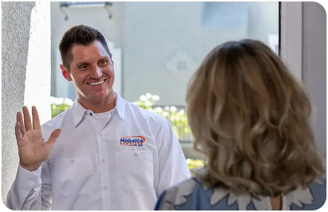 A man in a Hobalca uniform smiles and waves while greeting a woman at her front door.
