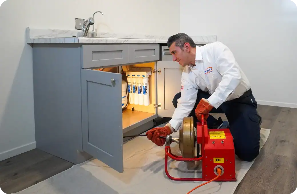 A plumber wearing gloves uses a drain cleaning machine next to a sink cabinet with filter cartridges visible inside.